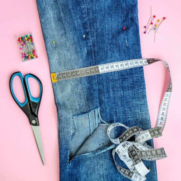 istock Folded in half blue jeans with large hole on pant leg, multi coloured headed sewing pins, white tailor tape with centimeters and inches and scissors. square image. 1157323354