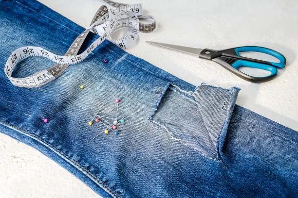istock Folded in half blue jeans with a large hole on pant leg below the knee, multi coloured headed sewing pins, white tailor tape with centimeters and inches and scissors 1157323358