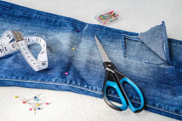 istock Folded in half blue jeans with large hole on pant leg below the knee, multi coloured headed sewing pins, white tailor tape with centimeters and inches and scissors. 1157323615