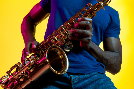 Young african-american jazz musician playing the saxophone on yellow studio background in trendy neon light. Concept of music, hobby. Joyful attractive guy improvising. Colorful portrait of artist.