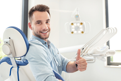 Handsome man showing thumbs up and smiling sitting at the dental chair. Professional dental clinic, healthy teeth concept.