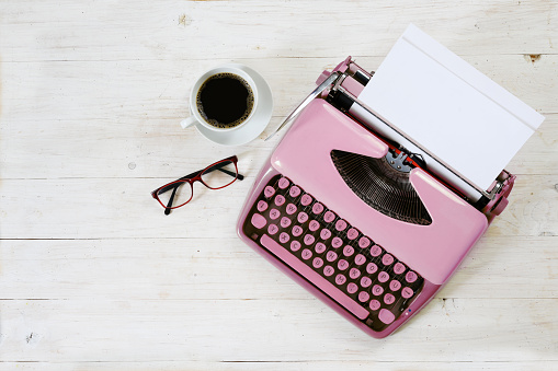 Old pink typewriter from the 1950s with blank paper, coffee and glasses on white painted rustic wood, copy space, high angle view from above, selected focus, narrow depth of field
