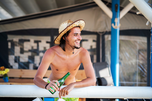 Young man with sun hat leaning on porch in campsite, he holding green beer bottle. looking away and smiling.