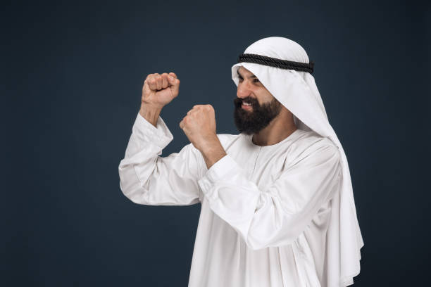 Portrait of arabian saudi businessman on dark blue studio background. Young male model standing, smiling and celebrating. Concept of business, finance, facial expression, human emotions.