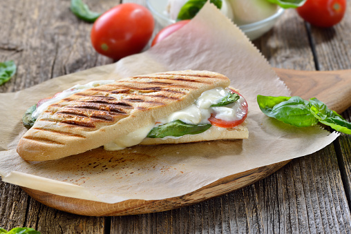 Pressed and toasted panini caprese with tomato, melted mozzarella and basil, served on sandwich paper on a wooden table