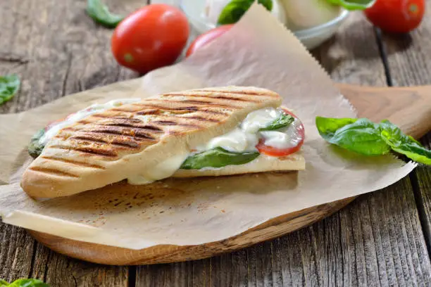 Pressed and toasted panini caprese with tomato, melted mozzarella and basil, served on sandwich paper on a wooden table