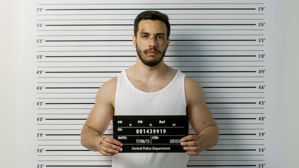 In a Police Station Arrested Beaten Man Poses for Front View Mugshot. He Wears Singlet, is Heavily Bruised and Holds Placard. Height Chart in the Background. In a Police Station Arrested Beaten Man Poses for Front View Mugshot. He Wears Singlet, is Heavily Bruised and Holds Placard. Height Chart in the Background. sleeveless top stock pictures, royalty-free photos & images