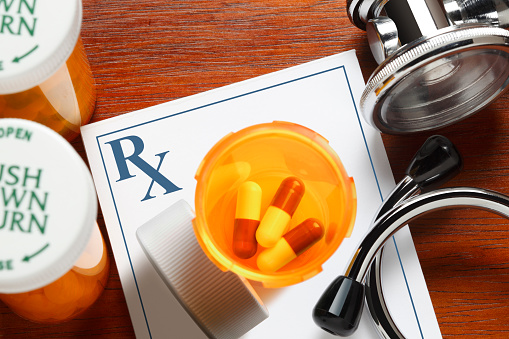 An open prescription medication pill bottle rests on top of a doctor's prescription pad and next to a stethoscope and other medication pill bottles.