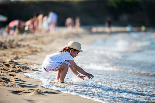 Little cute girl wearing dress and sun hat touching the sea on the summer beach.