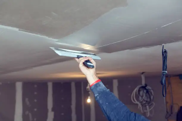 Worker is working with palette-knife for flattening  the ceiling from wooden platform in room of  apartment is inder construction, remodeling, renovation, overhaul, extension, restoration and reconstruction. Concept of home improvement