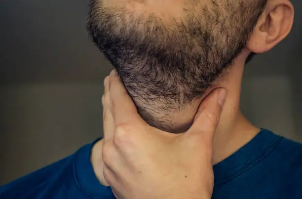 A young man with a sore throat. With his hand on the neck, sharp pain in the throat. Close up.