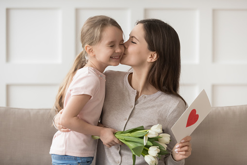 Mom kissing little daughter on cheek express gratitude for white tulips and drawn red heart on postcard symbol of devotion and love. Mother day eight march international womens day celebration concept