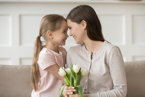 Cheerful mommy holds white tulips bouquet of flowers touches foreheads with daughter sitting on couch, little kid congratulate mom with spring holiday of 8 march, mother day or happy birthday concept