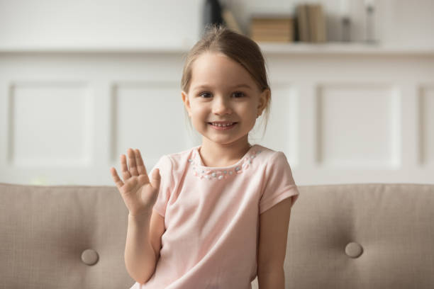 Headshot portrait little cheerful girl wave hand looking at camera Head shot portrait little girl sitting on sofa at home wave hand look at camera use webcam online communication with friend, relative, grandparents, kid greeting make video call recording vlog concept kid waves stock pictures, royalty-free photos & images