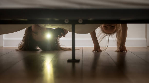 Mother and daughter shine a flashlight looking under the bed Mother and daughter, babysitter and kid girl afraid of monsters hold smartphone shine a flashlight under the bed looking on floor check ghosts, older younger sisters playing having fun at home concept nanny photos stock pictures, royalty-free photos & images