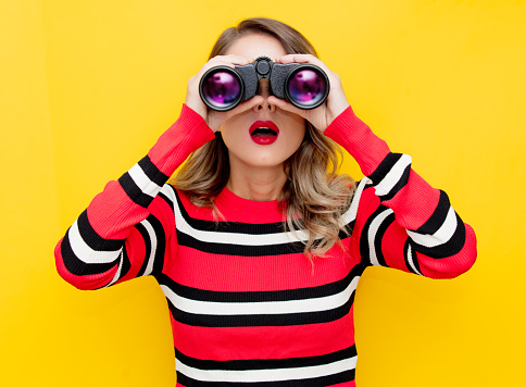 young woman in striped sweater with binocular on yellow background