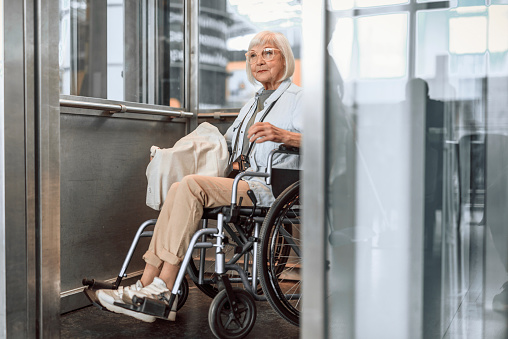Full length portrait of old female holding white bag and sitting in wheelchair while taking lift