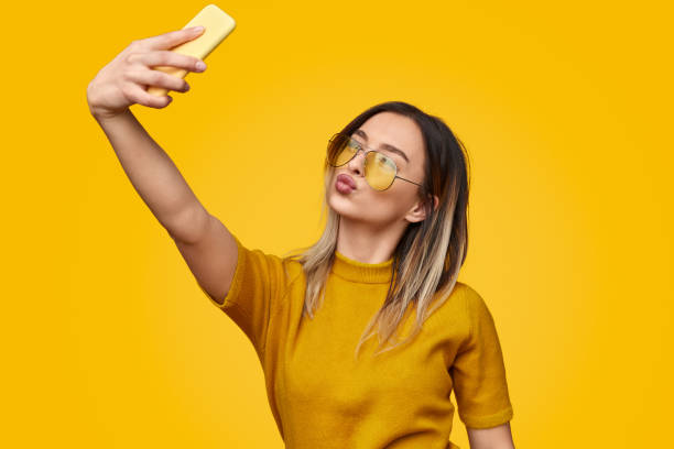 Stylish female pouting lips and taking selfie Young woman in trendy outfit pouting lips and using smartphone to take selfie while standing against yellow background photo messaging stock pictures, royalty-free photos & images