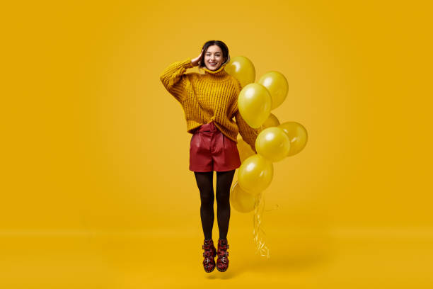 Weird female with balloons jumping and saluting Full body freakish young lady in colorful outfit holding bunch of vivid balloons and saluting while jumping against yellow background eccentric stock pictures, royalty-free photos & images