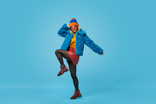 Full length weird young lady in colorful stylish outfit lifting leg and touching hat while dancing against blue background