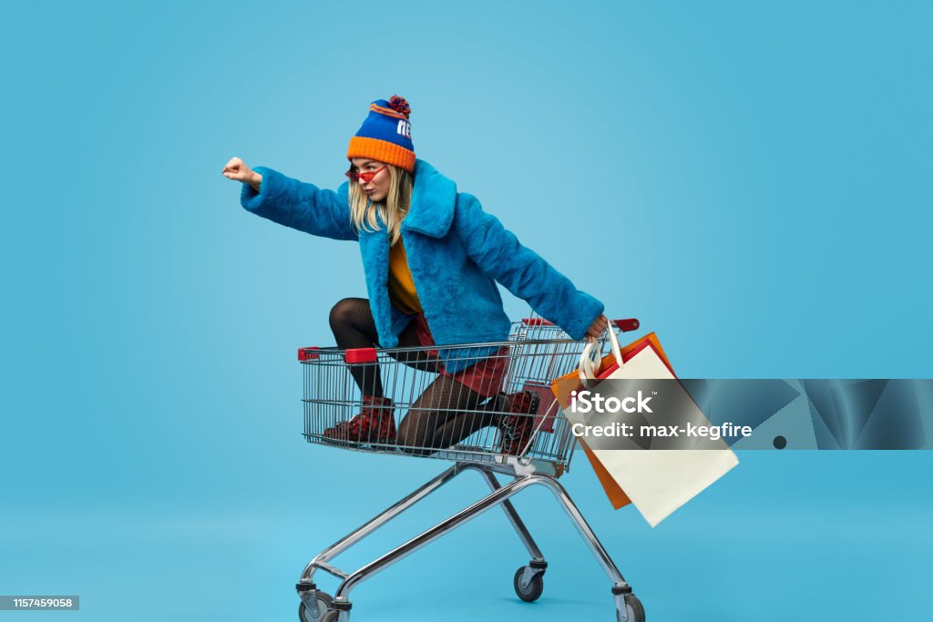 Young woman with shopping bags riding trolley Side view of crazy trendy young female in bright clothes holding paper bags and riding shopping cart in superhero pose against blue background Retail Stock Photo