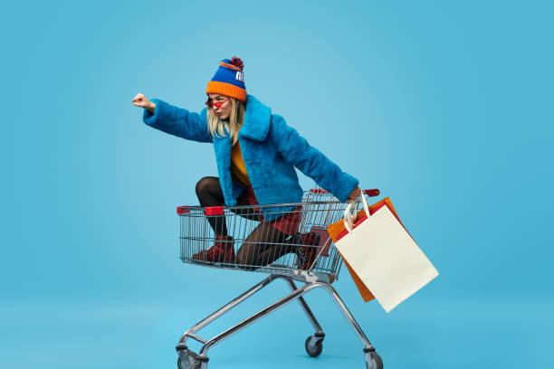 Young woman with shopping bags riding trolley Side view of crazy trendy young female in bright clothes holding paper bags and riding shopping cart in superhero pose against blue background bizarre stock pictures, royalty-free photos & images