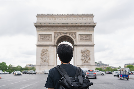 a man with backpack looking at Arc de Triomphe, famous landmark and travel destination in Paris, France. Traveling in Europe in summer