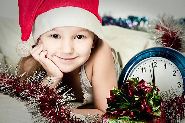 Adorable caucasian girl in christmas Santa hat with alarm clock in her hands. A place for your text. Studio shot on a yellow background. The concept of New Year and Christmas.