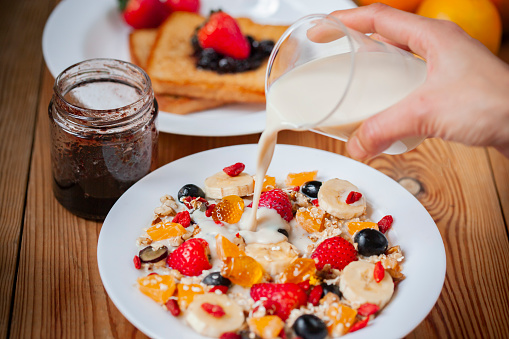 Table top, close-up image of hands pouring soy milk on healthy homemade oats breakfast with jam bread and various fruit toppings in white plate on wooden dining table.