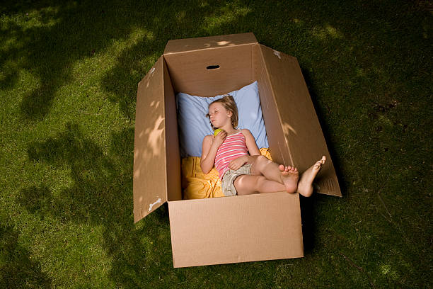 Young girl asleep Under a Tree in brown cardboard box.