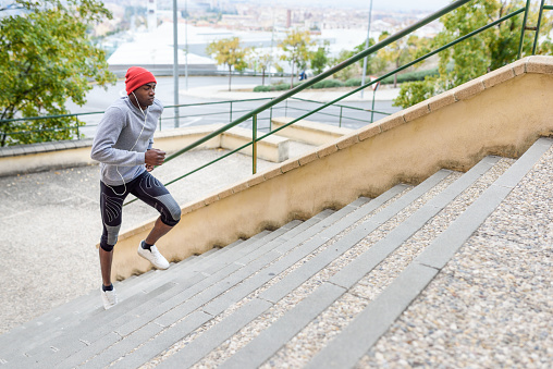 Black man running upstairs outdoors listening to music with white headphones. Young male exercising with city scape at the background.