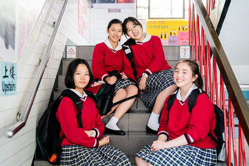 Portrait of smiling female high school students sitting on steps. Schoolgirls are enjoying in school building. They are wearing red uniforms.