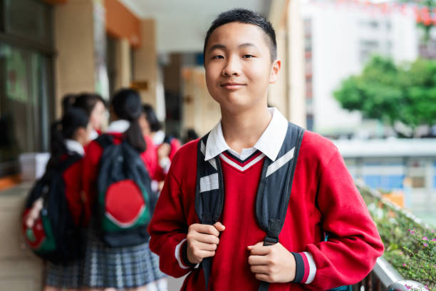 Portrait of confident teenage schoolboy in campus Portrait of confident teenage schoolboy holding backpack strap. Male high school student is standing in corridor. He is wearing red sweater. boy and girl looking at camera stock pictures, royalty-free photos & images