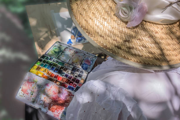 Woman artist in a beautiful straw hat, decorated with white feathers and flowers, sits in front of the easel. The girl sits back to us in a white lace vintage dress. Artistic background.