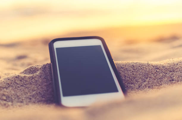 istock Smart phone on tropical sunset beach with bokeh light sand texture background. Black copy space of technology and travel nature concept. Vintage tone filter effect color style. 1157500124