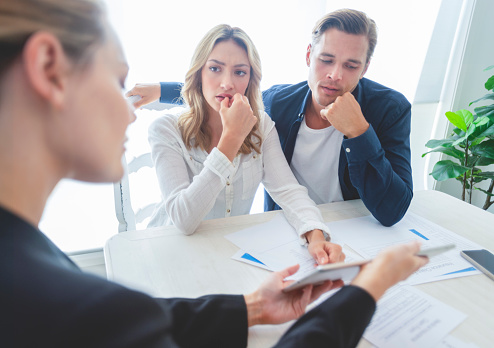 Real estate agent with couple looking through documents. The agent is holding a digital tablet showing it to the clients. Couple are casually dressed. They are looking concerned and worried and upset. Over the shoulder view.