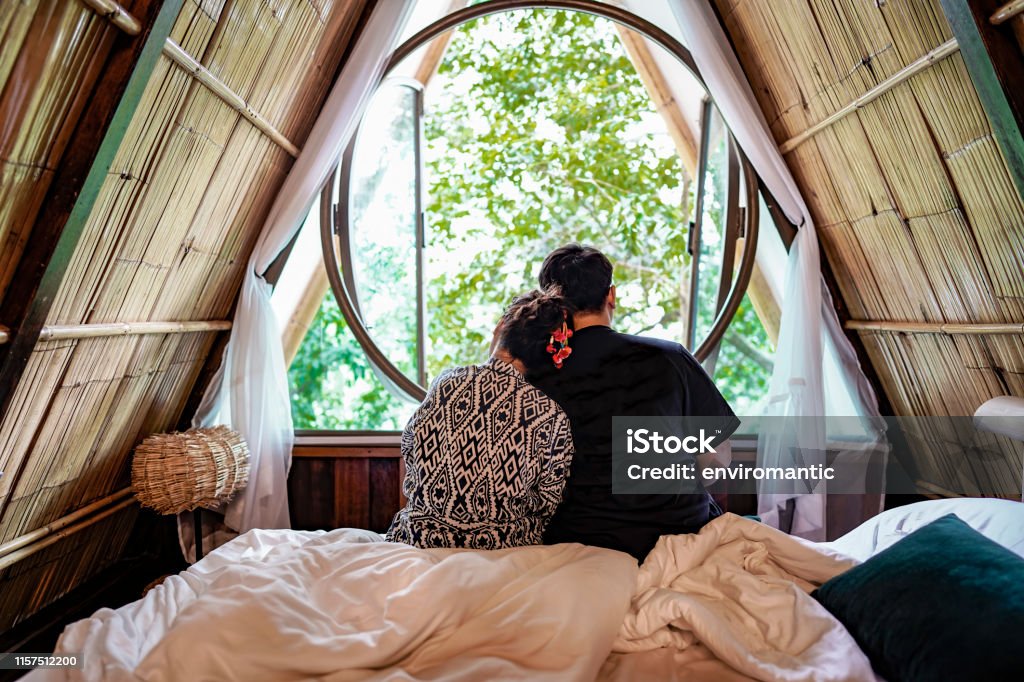 Young couple sit close together admiring nature looking out though a window of a rustic, natural guest house bedroom. Young loving couple sitting close together reflecting on the natural scene of trees in nature looking out though a window of a rustic, natural guest house bedroom. Bamboo - Material Stock Photo