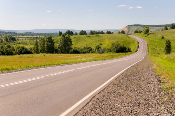 istock Asphalt road going across mountains and green forests. Trees and their shadows on the grass. Sunny summer day with blue sky. Ural landscape 1157538357