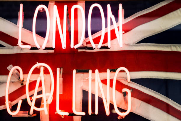 Close up image of an illuminated neon sign spelling out the words 'London Calling' in reference to the song by The Clash of the same name. Behind the neon words is the British flag - the Union Jack - in its famous red, white and blue. Horizontal colour image with copy space.