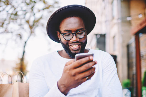 istock Smiling dark skinned guy in trendy hat read information about delivery service on web page using mobile phone, positive african american male satisfied with online shopping sales browsing on cellular 1157540374