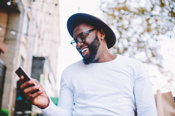istock Cheerful African American man in white shirt using mobile phone application for searching cafes near location in downtown, happy dark skinned hipster guy read news from social network walking in city 1157540383