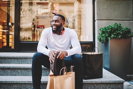 Smiling african american male customer in trendy wear sitting on stairs of store with bags with copy space for label, cheerful dark skinned hipster guy recreating after shopping and buying purchases