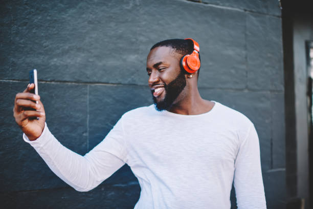 istock Cheerful african american guy in headphones spending time outdoors with modern gadgets taking picture on smartphone camera, smiling dark-skinned male hipster making selfie while listening music 1157541157