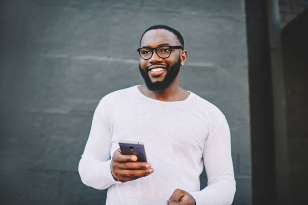 istock Cheerful african american male in spectacles using mobile phone outdoors looking waya and smiling, positive dark skinned hipster guy in white shirt with copy space for brandname holding cellular 1157541164