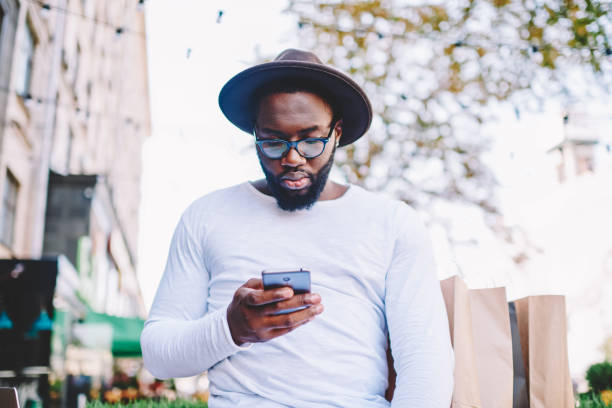 istock Pensive african american hipster guy in trendy hat installing application on smartphone spending time outdoors,thoughtful dark skinned man in shirt with copy space for brand name reading news on phone 1157541171
