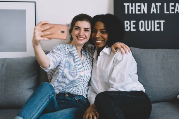 istock Two happy multicultural bloggers hugging each other and smiling at front camera making selfie on smartphone.Cheerful friends resting on cozy couch and taking pictures for publication on website 1157543191