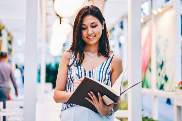 istock Smiling young female organizing and planning  information making notes while standing outdoors, attractive asian girl drawing in sketchbook enjoying free time on hobby making creative graphic 1157550119
