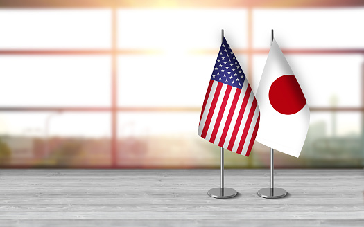 3D USA and japanese flags with metallic pole, standing together on a white wooden desk in front of sunny window background. With large copy space you can write your own titles effectively. Also you can use this compositon as square in social media channels like instagram etc.
