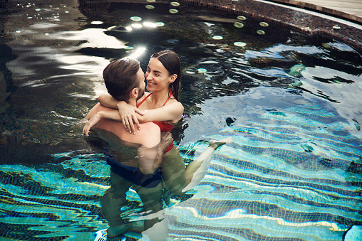 Man dearly holding black-haired woman in red swimsuit while spending time in the pool outdoors