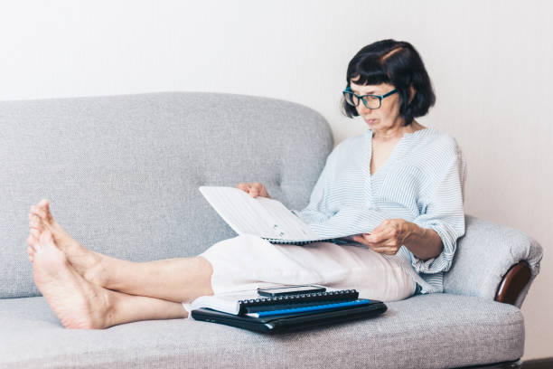 istock Middle-aged brunette woman on the gray sofa reading documents, using laptop and smartphone 1157599905